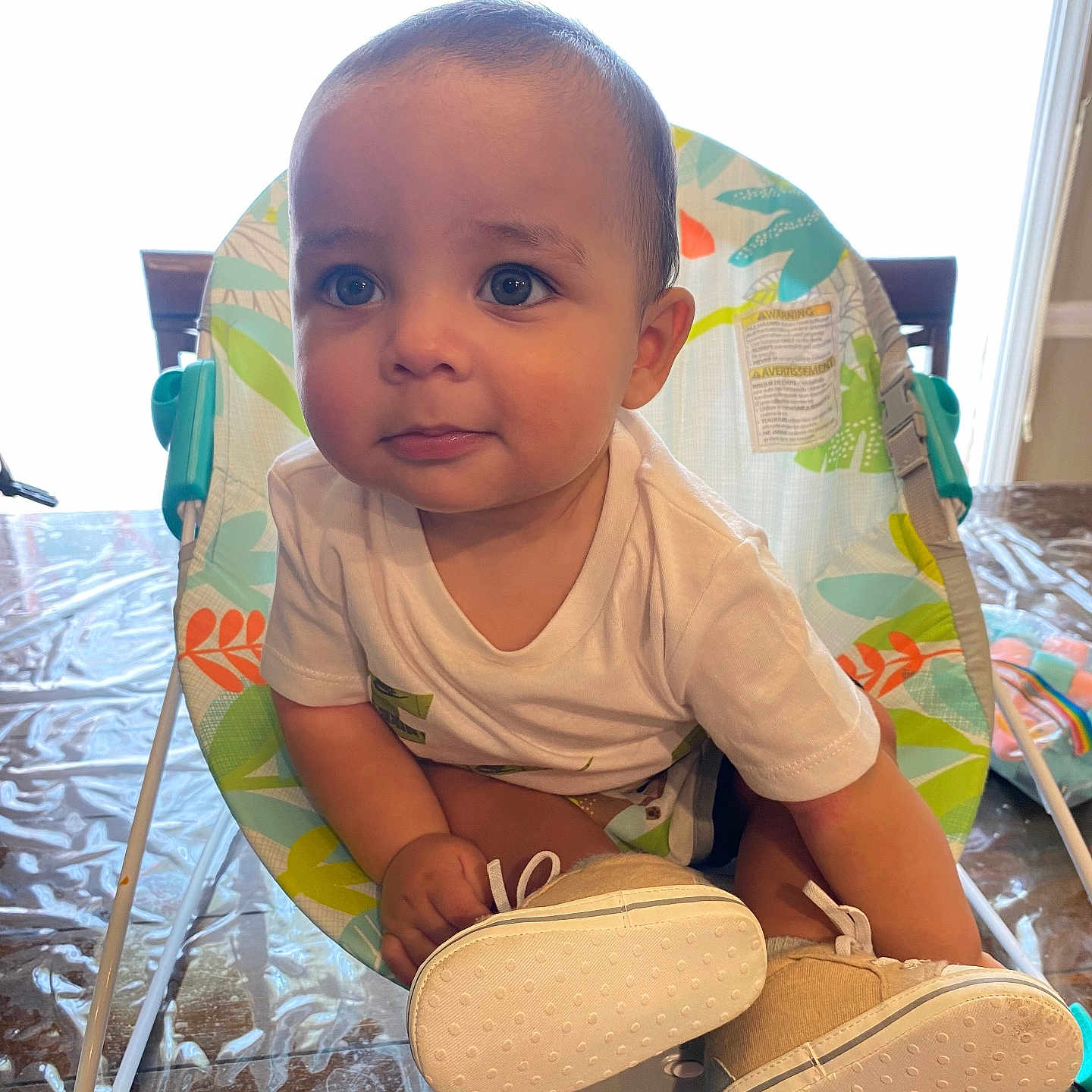 Noah is registered to the contest to win money with this photo: baby, baby_chair, bright, child, curious, cute, expression, eyes, face, footwear, furniture, indoor, infant, person, plastic_cover, shirt, shoes, sitting, window, wooden_table