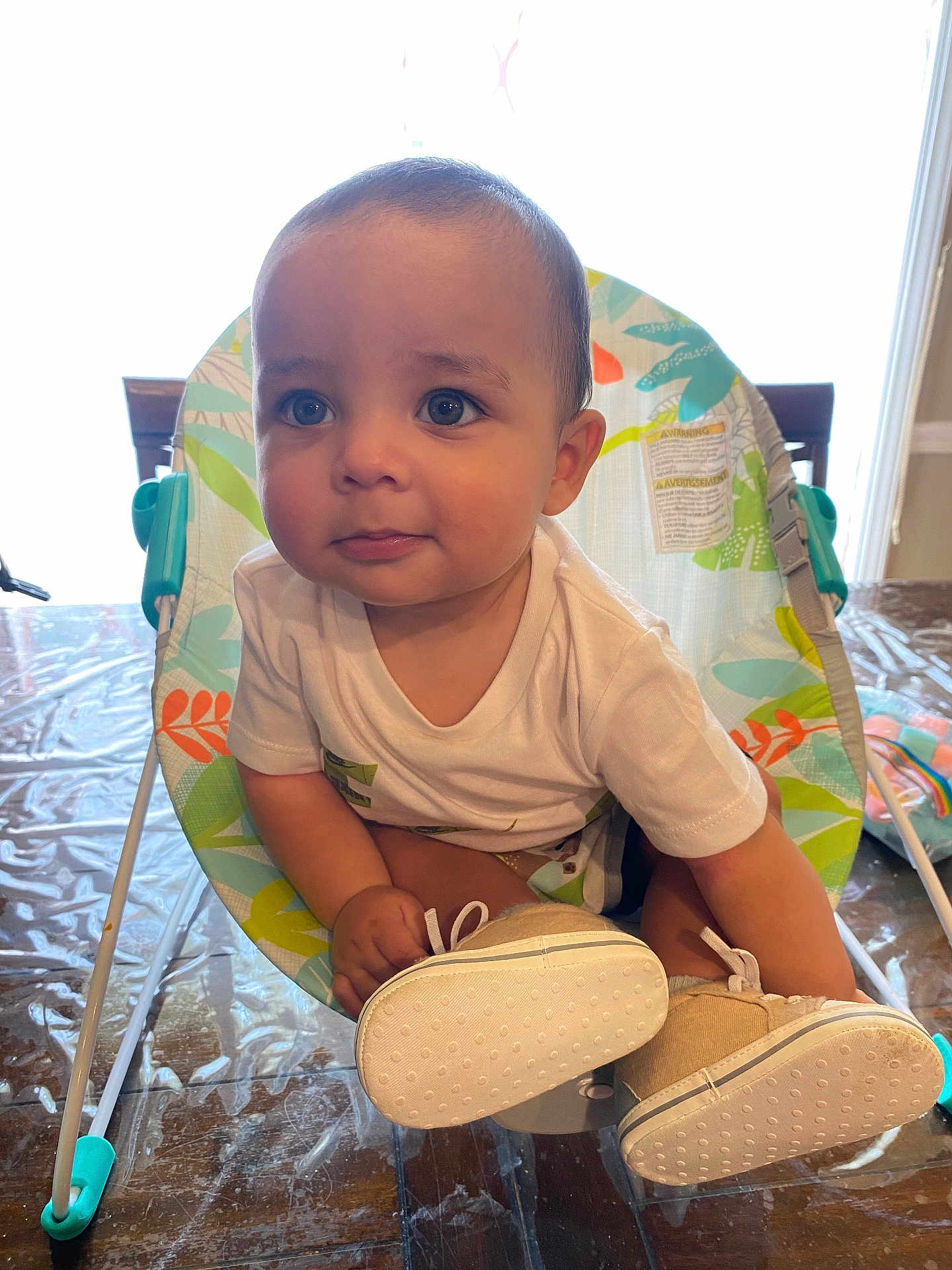 Noah is registered to the contest to win money with this photo: baby, child, infant, face, eyes, shirt, shoes, baby_chair, wooden_table, plastic_cover, indoor, curious, cute, person, sitting, footwear, expression, bright, window, furniture