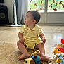 toddler, child, yellow_clothing, sitting, toy, stuffed_animal, floor, indoor, fan, speaker, window, door, sticker, natural_light, curious, looking_away, barefoot, home, playtime, colorful