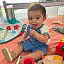 baby, child, blue_overalls, toy, blanket, indoor, play, smiling, tongue_out, window, natural_light, red_toy, sandals, fabric_toy, colorful, cute, happy, person, floor_play, infant
