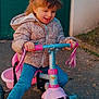child, girl, tricycle, outdoor, smiling, curly_hair, jacket, blue_jeans, toy, play, happy, daylight, pavement, green_door, pink, pedal, handlebar, casual_clothing, fun, recreation