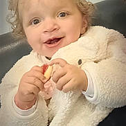 Zaina a rejoint le concours — aidez-le/la à gagner de superbes lots ! child, toddler, curly_hair, smiling, indoor, white_jacket, snack, cute, happy, face, person, cozy, seated, baby, small_hands, light_skin, clothing, portrait, candid, joyful