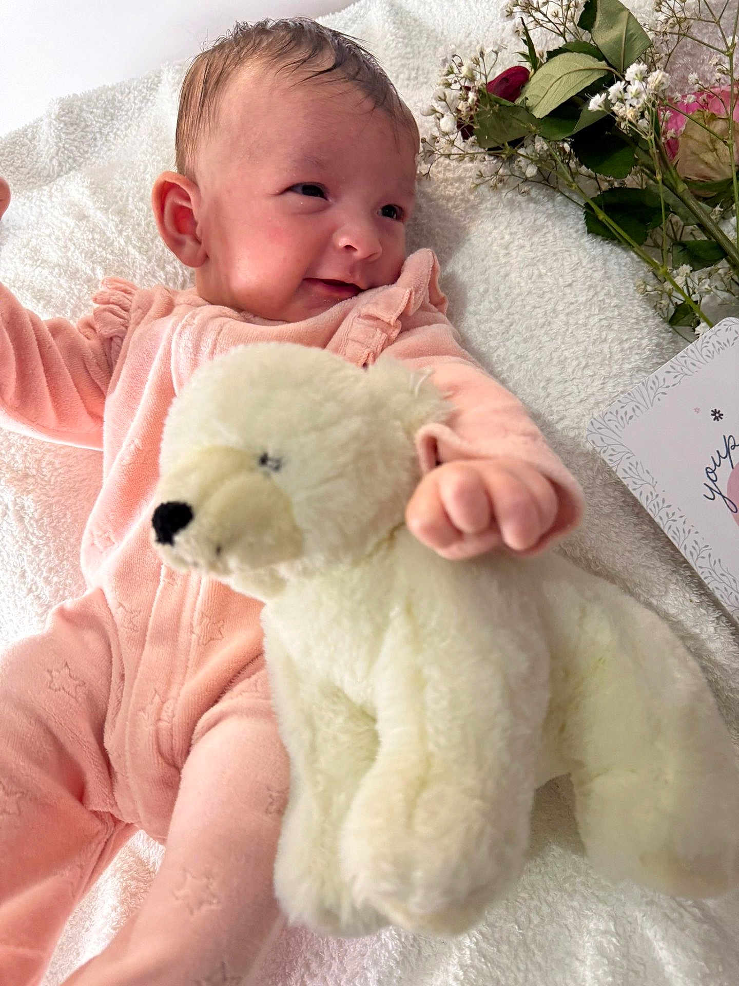 Inaya participe au concours pour gagner de l'argent avec cette photo : baby, infant, pink_onesie, stuffed_animal, white_bear, blanket, flower, card, smiling, cute, soft_toy, newborn, child, lying_down, hand, face, portrait, cozy, comfort, happy