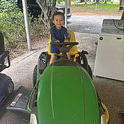 Raylon joined the competition — help win amazing prizes! child, toddler, smiling, lawn_mower, green, outdoor, covered_area, trees, truck, washing_machine, grass, concrete_floor, yellow_seat, playful, vehicle, happy, person, daylight, nature, home