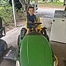 child, toddler, smiling, lawn_mower, green, outdoor, covered_area, trees, truck, washing_machine, grass, concrete_floor, yellow_seat, playful, vehicle, happy, person, daylight, nature, home