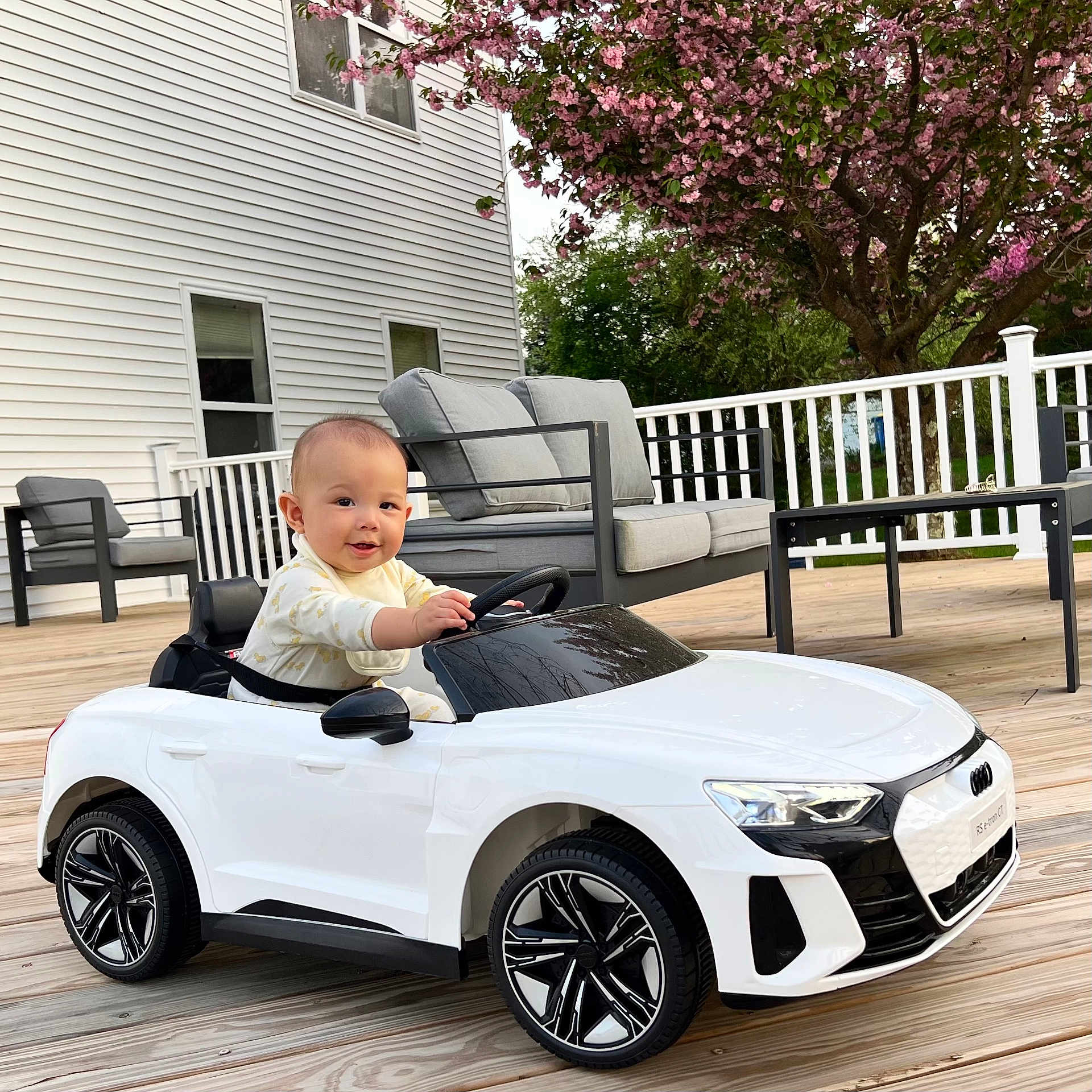 Joaquin is registered to the contest to win money with this photo: baby, baby_clothing, car_wheel, chair, child, cushion, deck, face, flowering_tree, house_exterior, outdoor, pink_flowers, seat, smiling, steering_wheel, table, toy_car, white_car, window, wooden_floor