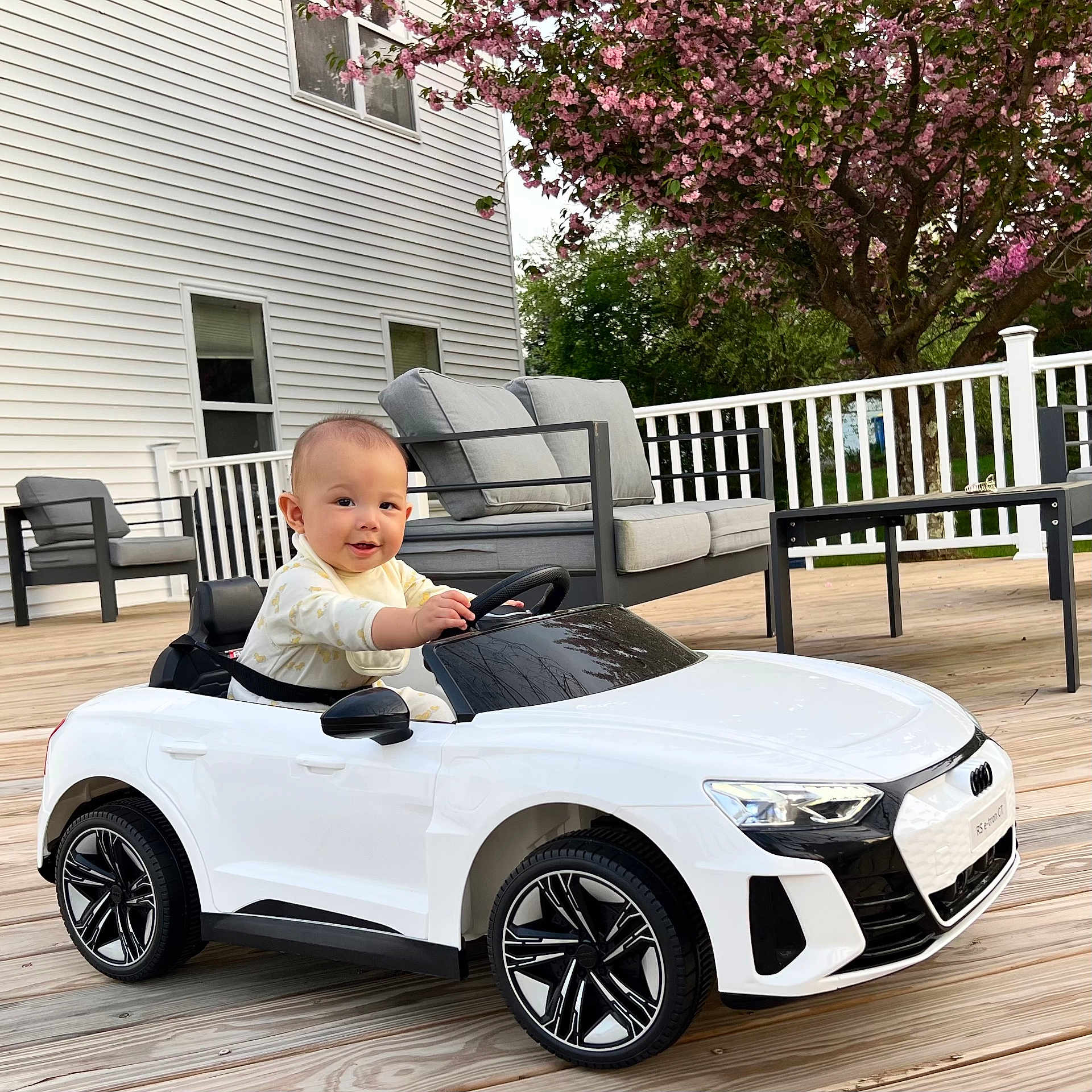Joaquin is registered to the contest to win money with this photo: baby, child, toy_car, white_car, outdoor, deck, wooden_floor, flowering_tree, pink_flowers, baby_clothing, steering_wheel, car_wheel, smiling, face, seat, cushion, table, chair, house_exterior, window