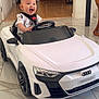baby, child, toy_car, indoor, smiling, happy, floor, fireplace, christmas_stockings, pumpkin, living_room, seatbelt, white_car, audi, head, person, small_vehicle, tile_floor, furniture, holiday_decor