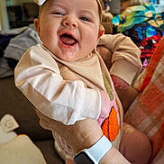 Emma is registered to the contest to win money with this photo: baby, child, smile, happy, face, headband, hand, watch, clothing, person, indoor, couch, colorful, background, skin, expression, holding, comfort, family, home