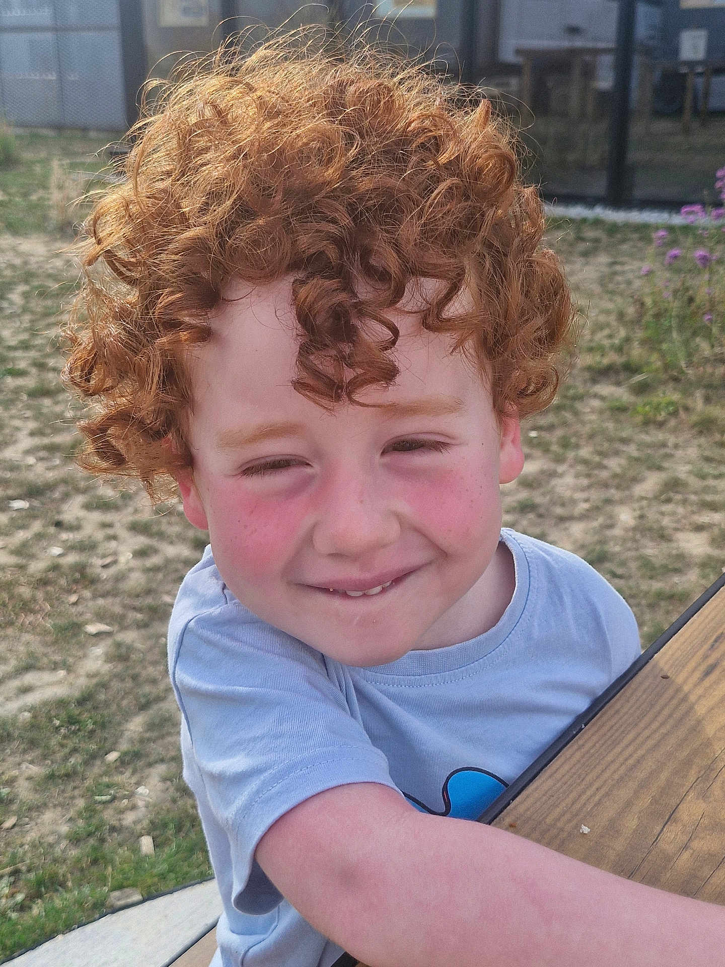 Maxen a rejoint le concours — aidez-le/la à gagner de superbes lots ! child, curly_hair, red_hair, smile, rosy_cheeks, outdoor, grass, wooden_table, tshirt, happy, face, person, nature, sunlight, freckles, cute, playful, young, boy, casual