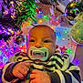 baby, pacifier, striped_clothing, christmas_tree, christmas_lights, ornaments, festive, holiday, colorful_lights, decorations, infant, closeup, cute, cozy, indoors, celebration, child, face, hands, winter
