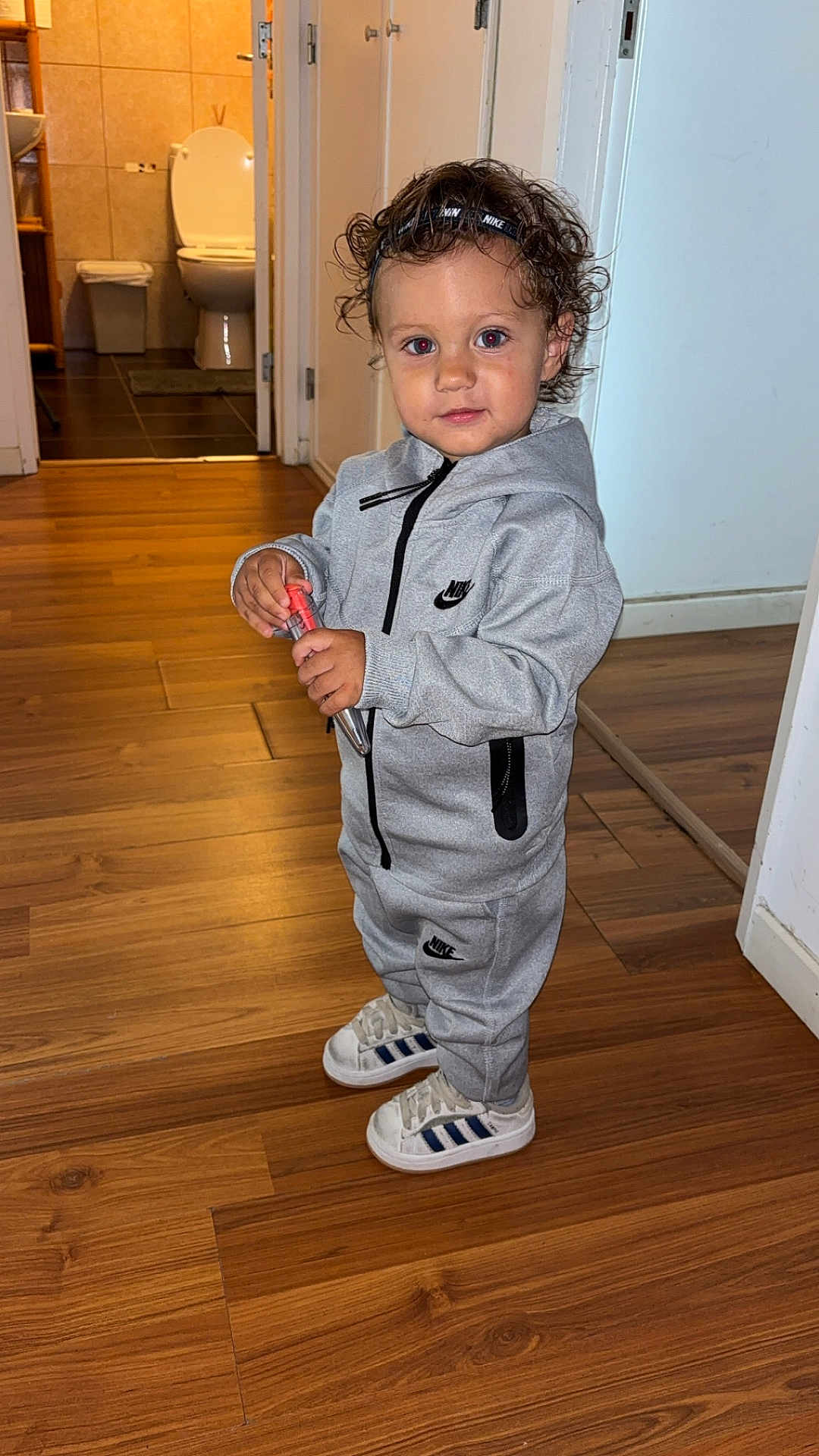 Lucas a rejoint le concours — aidez-le/la à gagner de superbes lots ! toddler, child, curly_hair, headband, nike, tracksuit, grey_clothing, sneakers, adidas, wooden_floor, indoor, person, standing, holding_object, bathroom, door, casual, home, flooring, portrait