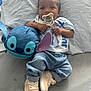 baby, infant, pacifier, plush_toy, stitch, denim_jeans, sneakers, white_shirt, bed, pillow, cute, child, lying_down, bracelet, indoor, soft_toy, casual_clothing, footwear, small_child, adorable