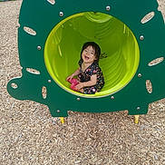 Anayelíz joined the competition — help win amazing prizes! toddler, child, playground, play_tunnel, smiling, happy, outdoor, grass, wood_chips, green, pink_shoes, floral_clothing, sitting, fun, daylight, play_equipment, nature, park, person, casual