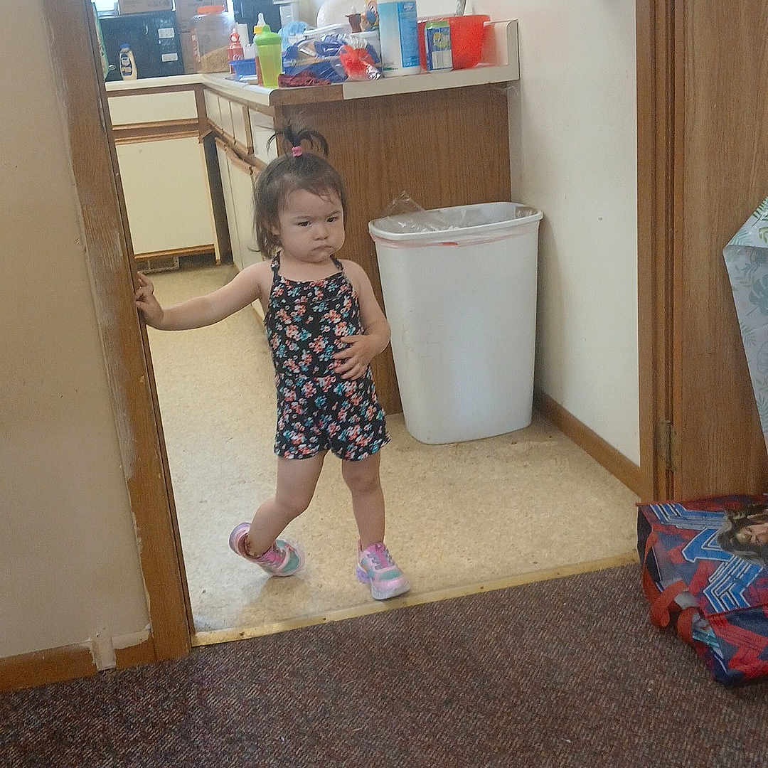 Anayelíz is registered to the contest to win money with this photo: box, carpet, child, counter, cup, doorway, fire_extinguisher, floor, girl, hair_tie, indoor, kitchen, plastic_bottle, romper, serious_expression, sneakers, standing, trash_can, wall, window