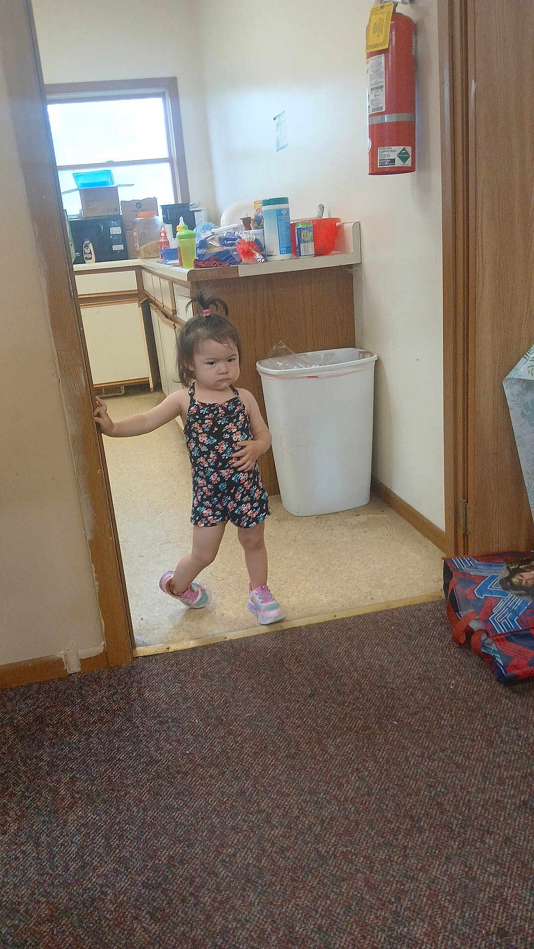 Anayelíz is registered to the contest to win money with this photo: child, girl, standing, doorway, romper, sneakers, hair_tie, kitchen, counter, trash_can, fire_extinguisher, carpet, floor, wall, window, plastic_bottle, box, cup, indoor, serious_expression