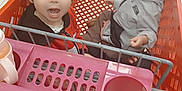 Misael is registered to the contest to win money with this photo: child, toddler, shopping_cart, store, indoor, person, clothing, pink_bow, floor, metal, plastic, sitting, face, expression, hair, young, retail, supermarket, toy, container