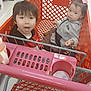 child, toddler, shopping_cart, store, indoor, person, clothing, pink_bow, floor, metal, plastic, sitting, face, expression, hair, young, retail, supermarket, toy, container