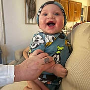 Messiah is registered to the contest to win money with this photo: baby, smiling, infant, cartoon_clothing, hat, hands, tattoo, home_interior, wooden_cabinets, warm_lighting, person, holding, joyful, cute, indoor, face, child, apparel, human, expression