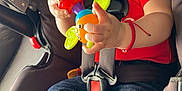 Anthony is registered to the contest to win money with this photo: baby, car_seat, pacifier, toy, child, red_shirt, blue_jeans, barefoot, straps, safety_harness, indoor, close_up, infant, expression, serious, colorful_toy, seat, foot, hand, person