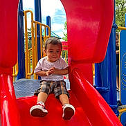 Arlo joined the competition — help win amazing prizes! child, toddler, slide, playground, red_slide, smiling, outdoor, summer, fun, clothing, shorts, white_shirt, sneakers, yellow_railings, blue_poles, play_structure, grass, trees, sky, happy