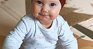 Joy a rejoint le concours — aidez-le/la à gagner de superbes lots ! baby, blue_eyes, brown_hair, child, chubby_cheeks, closeup, cute, floor_mat, hands, headband, indoor, infant, onesie, play_mat, portrait, sitting, smile, soft_lighting, tiled_floor, toy_ball