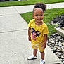 child, toddler, smiling, curly_hair, yellow_shirt, yellow_shorts, sneakers, sidewalk, outdoor, green_grass, rocks, casual_clothing, happy, person, portrait, daylight, urban, playful, cute, parked_car