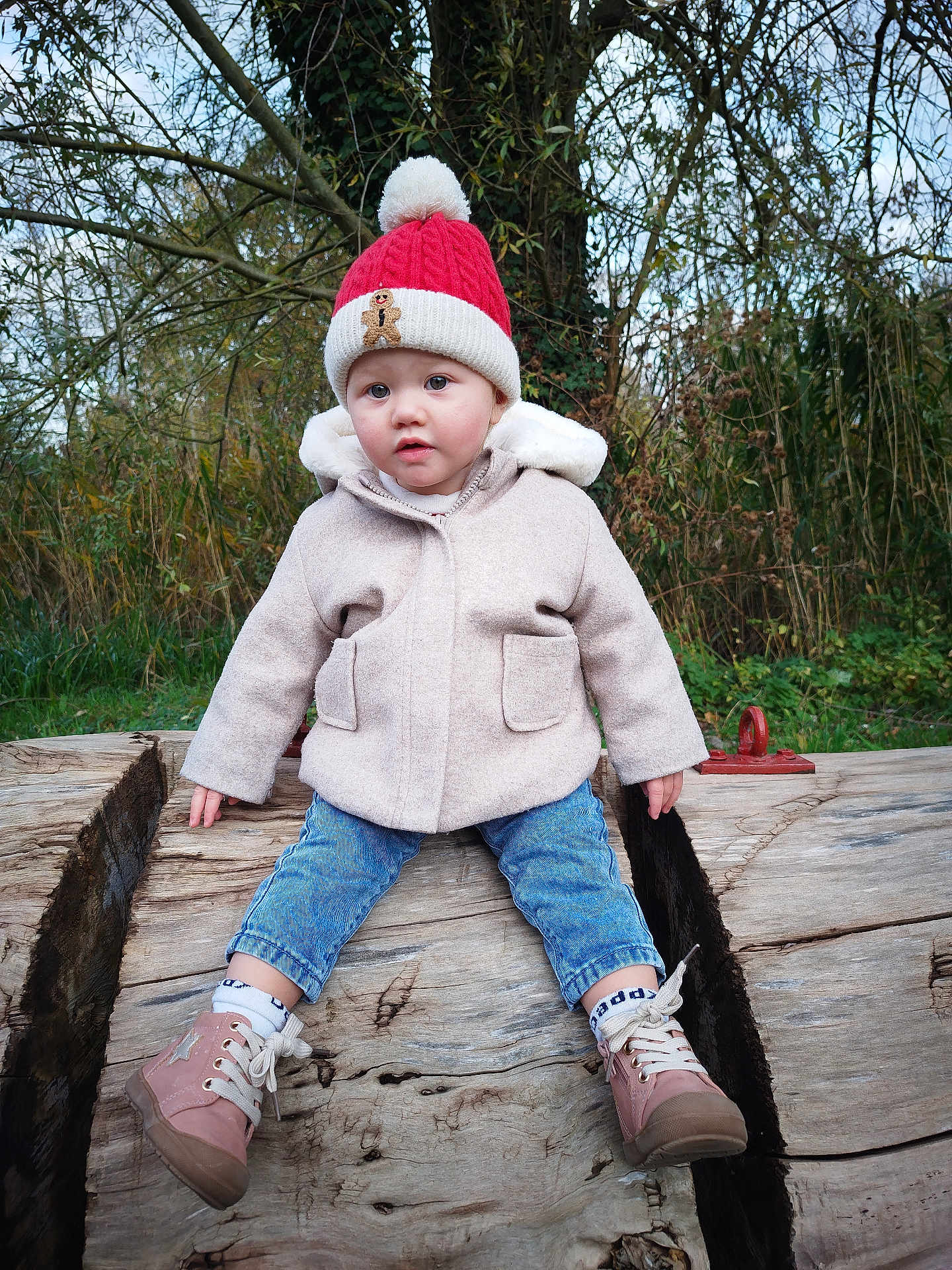 Elyana participe au concours pour gagner de l'argent avec cette photo : toddler, child, outdoor, winter_clothing, hat, pom_pom_hat, coat, jeans, boots, log, tree, forest, nature, grass, face, person, sitting, curious, cute, cold_weather