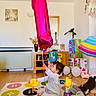 balloon, balloons, birthday, celebration, child, colorful_rug, curly_hair, decorations, furniture, gift_box, home, indoor, mirror, number_one, person, play, smiling, toddler, toy, wooden_floor