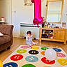 baby, balloon, child, colorful_rug, curly_hair, curtains, decor, floor, furniture, home, indoor, living_room, mirror, number_one, plastic_container, playful, toddler, toy, white_balloon, wooden_cabinet
