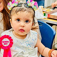Huda is registered to the contest to win money with this photo: birthday, bow, bracelet, celebration, chair, child, cute, dress, eyes, face, festive, food, girl, hair_accessories, indoor, party, people, ribbon, seated, table