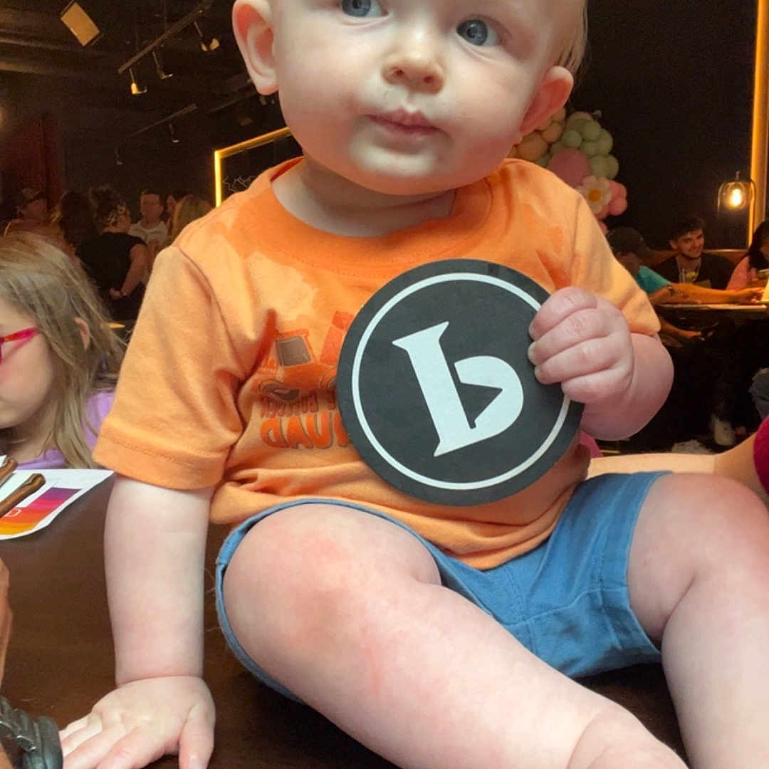 Landon is registered to the contest to win money with this photo: accessories, baby, bodypart, clothing, face, finger, furniture, glasses, hand, head, indoors, pants, person, photography, portrait, shorts, sitting, t_shirt, table, wood