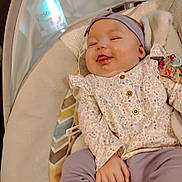 Emberlee is registered to the contest to win money with this photo: baby, infant, smile, headband, floral_clothing, purple_pants, baby_seat, cushion, hand, face, indoors, cozy, cute, playful, child, person, relaxing, comfort, soft_fabric, patterned_background