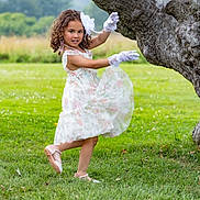 Aria is registered to the contest to win money with this photo: child, girl, dress, gloves, tree, grass, outdoor, nature, field, mountain, flower_pattern, curly_hair, pose, smile, footwear, greenery, playful, young, daylight, scenic