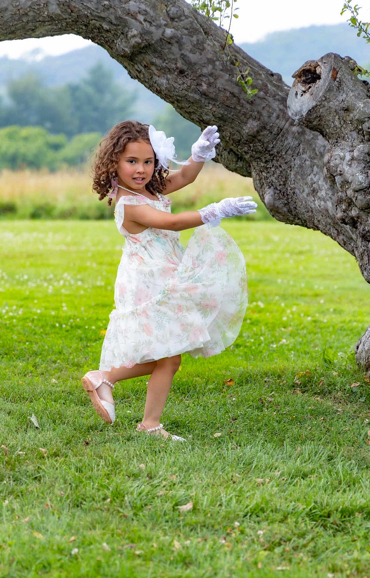 Aria is registered to the contest to win money with this photo: child, girl, dress, gloves, tree, grass, outdoor, nature, field, mountain, flower_pattern, curly_hair, pose, smile, footwear, greenery, playful, young, daylight, scenic