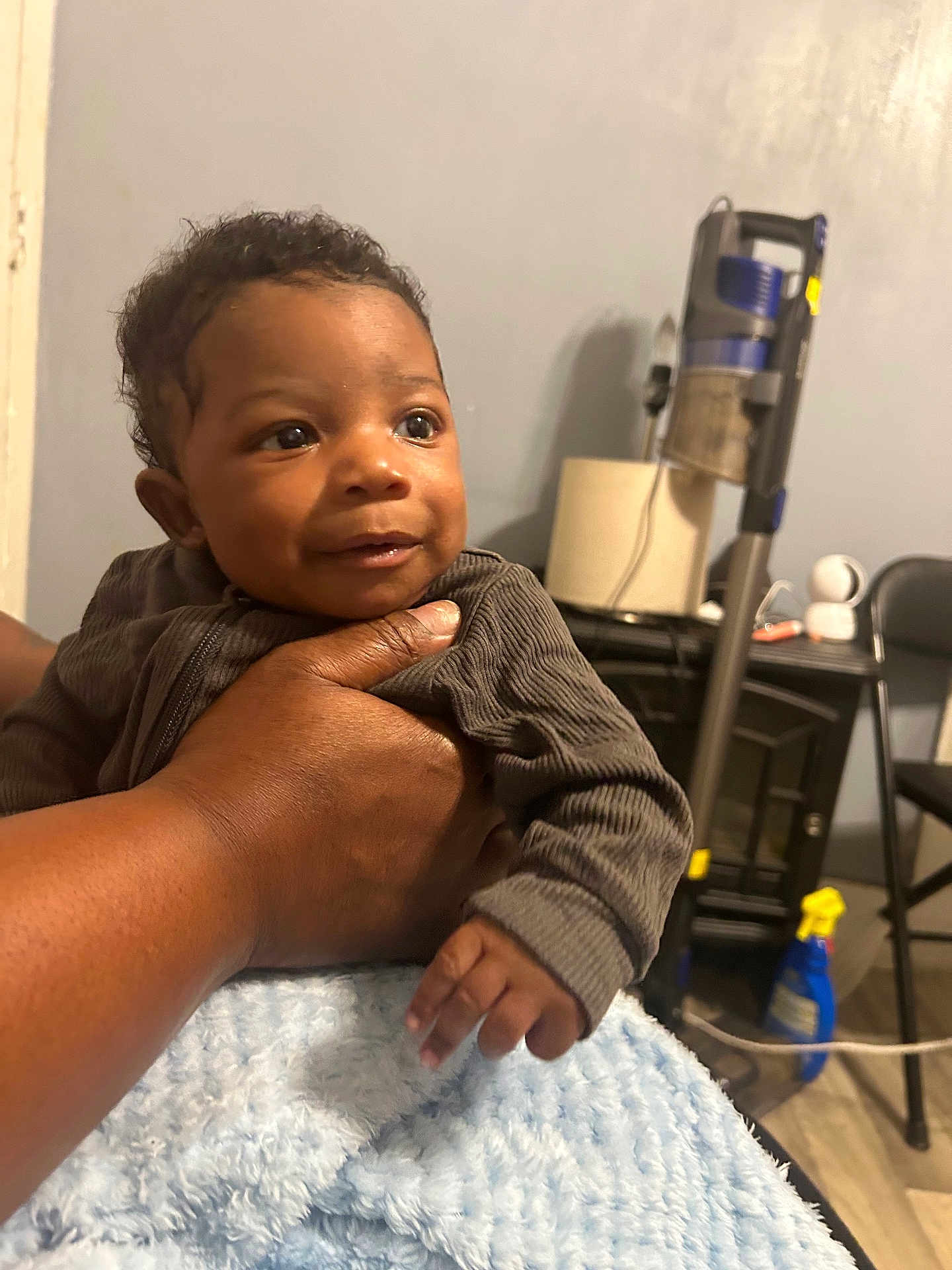 Zakari is registered to the contest to win money with this photo: baby, child, hand, blanket, indoor, person, furniture, vacuum_cleaner, spray_bottle, table, chair, wall, floor, clothing, smile, face, portrait, cozy, curly_hair, cute