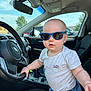 baby, child, sunglasses, car_interior, steering_wheel, seat, clothing, toddler, summer, daylight, window, sky, dashboard, cute, portrait, person, vehicle, seatbelt, casual, blue