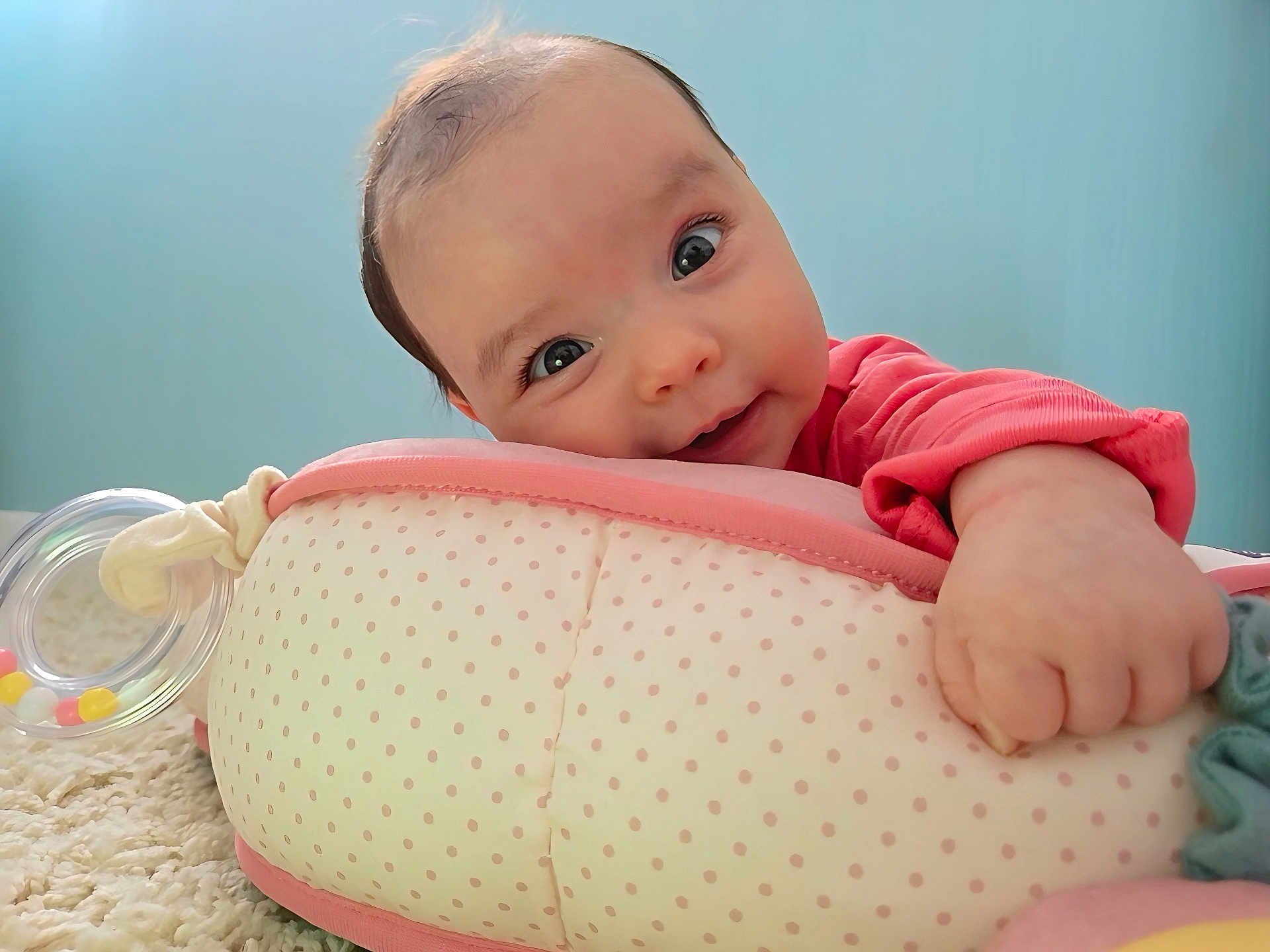 Mia joined the competition — help win amazing prizes! baby, infant, face, hand, pink_clothing, polka_dots, cushion, carpet, blue_wall, curious, grip, indoors, soft_texture, close_up, child, cute, portrait, young, smiling, baby_toy