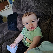 Kameron is registered to the contest to win money with this photo: baby, child, smiling, blue_eyes, green_shirt, white_socks, couch, blanket, plush, indoor, toy, happy, person, seated, infant, cute, comfortable, home, portrait, cozy