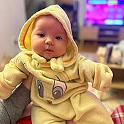 Vienna is registered to the contest to win money with this photo: baby, blanket, blurred_background, child, cozy, cute, face, fuzzy, hand, homedecor, hooded_clothing, indoor, infant, person, portrait, sitting, smiling, soft_texture, warm_clothing, yellow_onesie