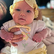 Vienna is registered to the contest to win money with this photo: baby, child, infant, yellow_bonnet, yellow_dress, clothing, cute, hands, person, face, portrait, indoor, soft_focus, skin, expression, newborn, holding, human, young, headwear
