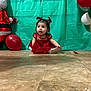 baby, balloons, celebration, child, curious_expression, cute, festive, floor, holiday, indoor, party_decoration, pigtails, plastic_backdrop, portrait, red_dress, santa_decoration, sitting, tile_floor, toddler, young_child