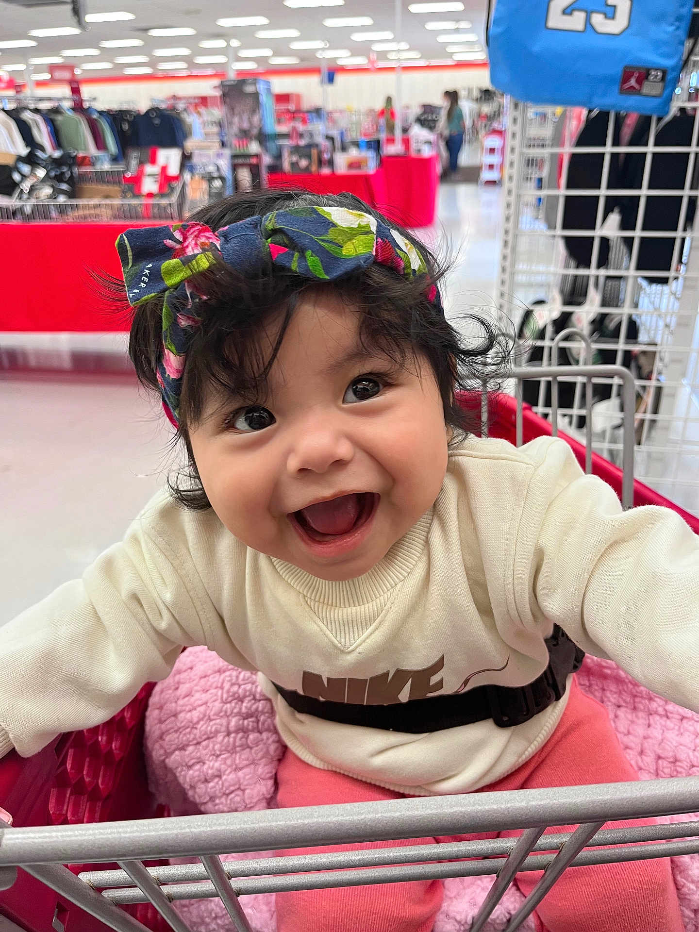 Melani joined the competition — help win amazing prizes! baby, child, smiling, shopping_cart, headband, clothing, indoor, store, retail, blanket, pink, sweater, happy, person, cute, face, curly_hair, bright_lighting, seatbelt, playful