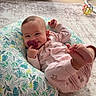 baby, infant, child, smiling, pacifier, pink_clothing, onesie, sitting, lying_down, barefoot, toes, cushion, patterned_cushion, rug, indoor, window, sunlight, houseplant, toy_ball, portrait