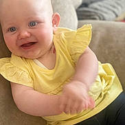 Arya is registered to the contest to win money with this photo: baby, child, smiling, yellow_top, blue_eyes, clapping, couch, indoor, happy, cute, sitting, person, infant, portrait, skin, head, hands, clothing, young, cheerful