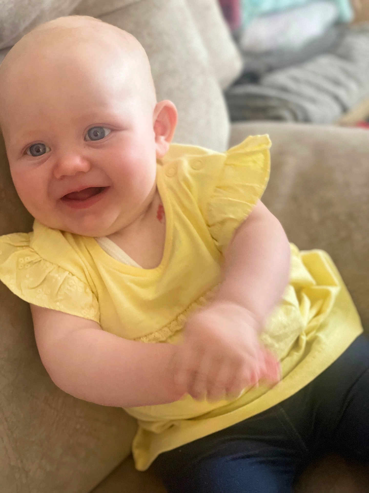 Arya is registered to the contest to win money with this photo: baby, child, smiling, yellow_top, blue_eyes, clapping, couch, indoor, happy, cute, sitting, person, infant, portrait, skin, head, hands, clothing, young, cheerful