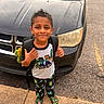 child, boy, smiling, backpack, minecraft, casual_clothing, black_car, parking_lot, sidewalk, footwear, green_shoes, outdoor, daylight, young_person, portrait, standing, short_hair, happy, vehicle, urban