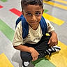 child, boy, backpack, sneakers, floor, colorful_floor, stripes, casual_clothing, crouching, looking_up, portrait, indoor, young, curious, smiling, short_hair, black_pants, white_shirt, shoe, person