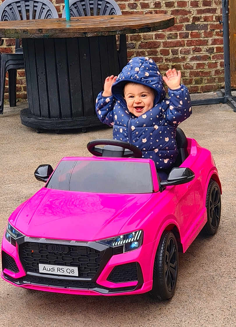 Camille participe au concours pour gagner de l'argent avec cette photo : toddler, child, toy_car, pink_car, hooded_jacket, blue_jacket, happy, smiling, outdoor, brick_wall, table, chair, concrete_ground, hands_up, play, vehicle, small_car, person, cute, joyful