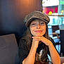 child, smile, hat, cap, long_hair, face, portrait, indoor, table, wood, clothing, person, seated, casual, young, happy, restaurant, blurred_background, fashion, hands